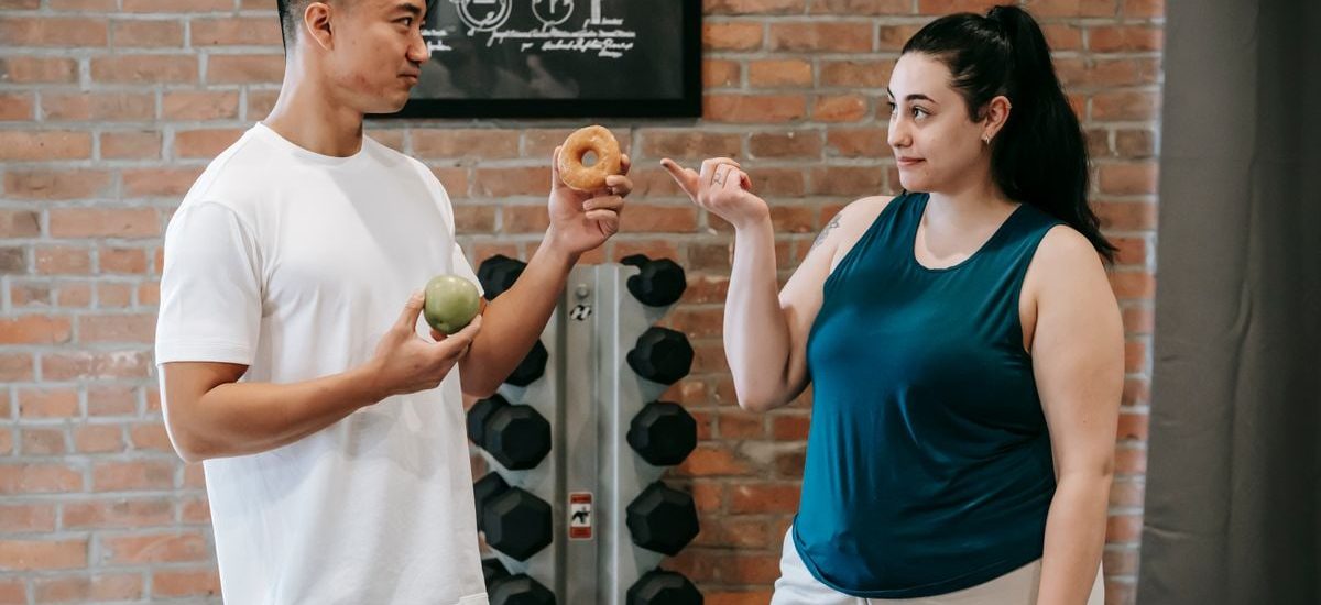 Side view of Asian personal instructor with apple and doughnut offering healthy food to plus size woman while standing in gym